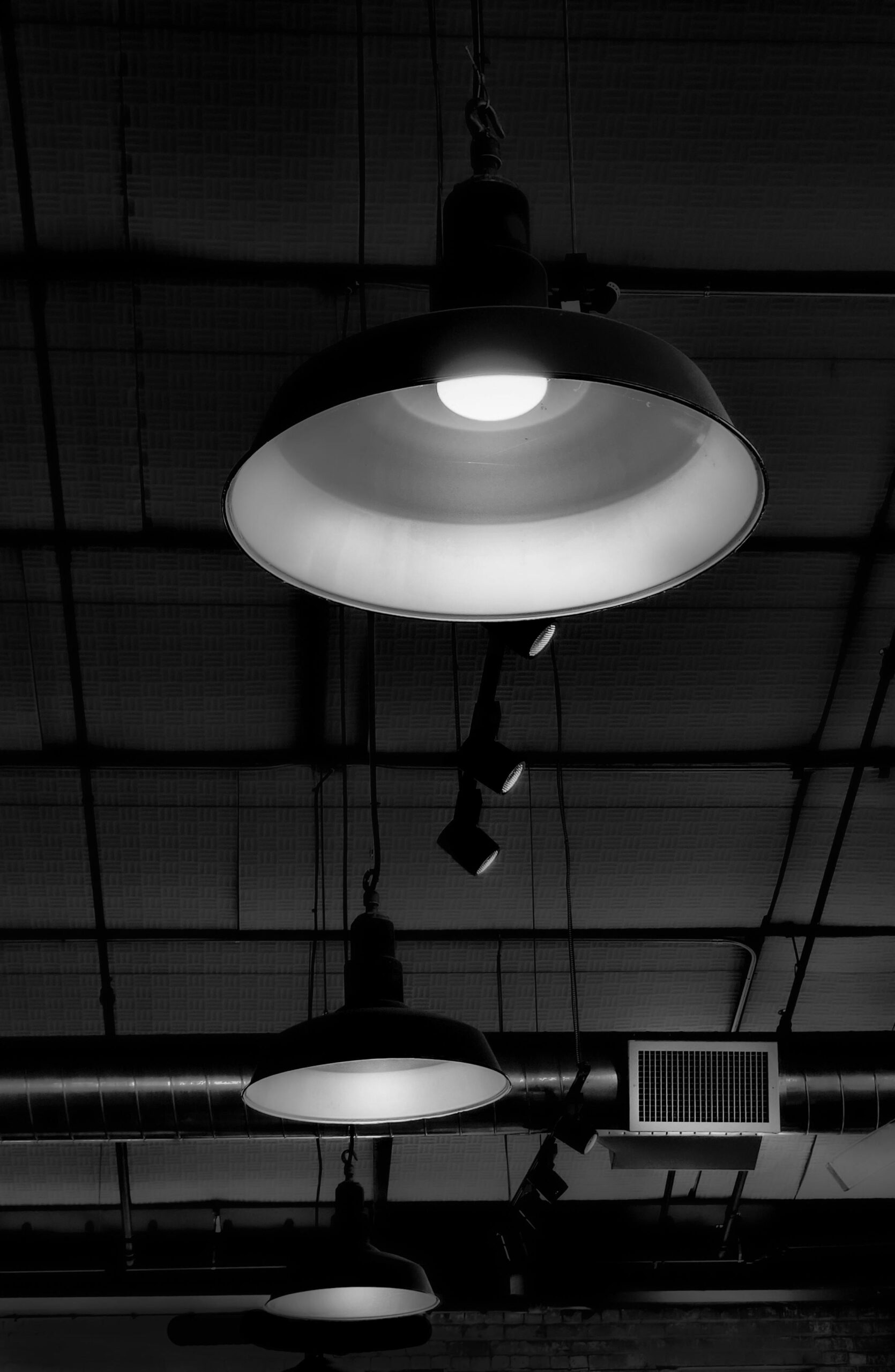 Black and white photo of industrial ceiling with hanging light fixtures, wires, and ductwork.